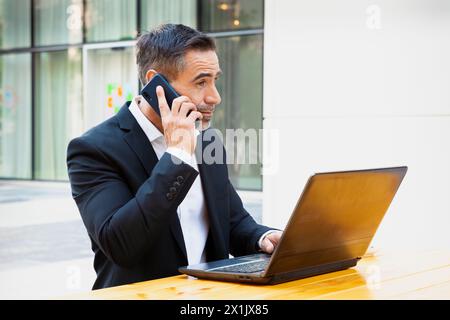 Businessman working with laptop and talking on smartphone smiling outdoors. Stock Photo