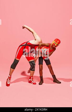 Two athletes in wrestling poses, man and boy in red outfit with mask against pink studio background. Fight. Stock Photo