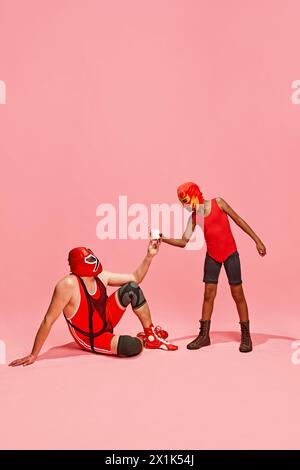 Boy in superhero dressed red costume gave glass of milk to his older, bigger partner against pink studio background. Stock Photo