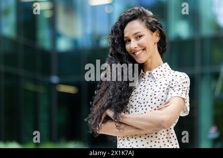A cheerful Hispanic woman with curly long hair smiles brightly standing against a modern glass building backdrop. She is wearing a polka dot shirt. Stock Photo