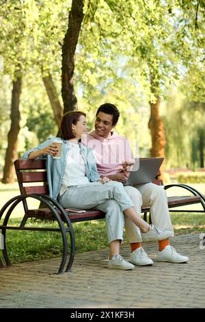 A stylishly dressed couple, immersed in the digital world, sits on a park bench, engrossed in the screen of a laptop. Stock Photo