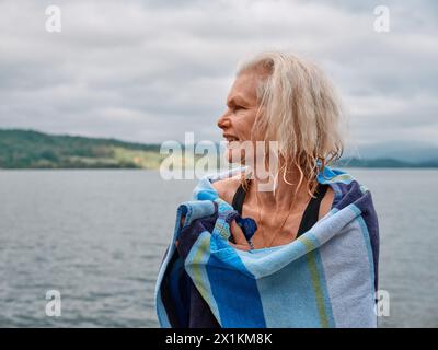 A senior open water wild swimmer warms themselves up with a towel after a refreshing cold water lake swim - woman swim outdoors health UK Stock Photo