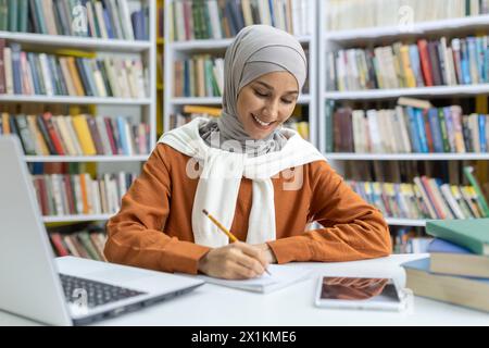 A cheerful young Muslim woman wearing a hijab studying in a library, surrounded by books. She is writing notes and using a laptop, exuding focus and happiness. Stock Photo