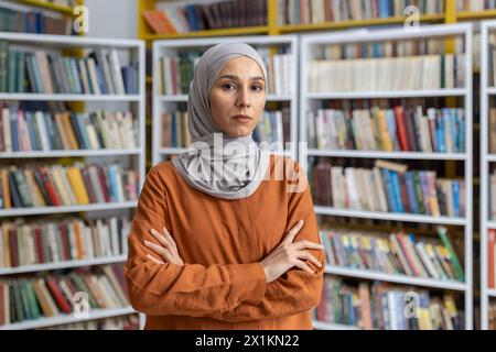 A confident Muslim woman in a hijab stands with her arms crossed in a library filled with books, portraying strength and intelligence. Stock Photo