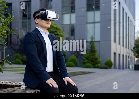 Serious and focused Asian male office worker in a business suit sitting on a bench near a building wearing a virtual mask. Stock Photo