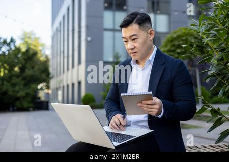 Serious young man, Asian businessman working outside an office building using a laptop and making notes in a notebook. Stock Photo