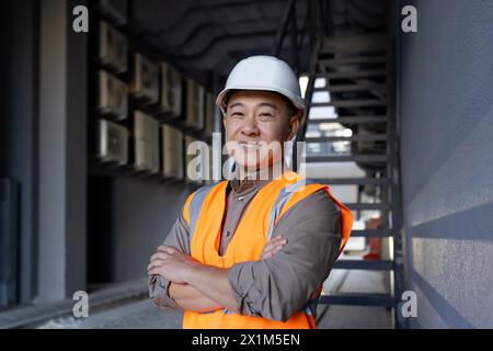 Portrait of a smiling Asian male engineer, construction worker, technician standing outside a building in a hard hat and vest, crossing his arms over his chest and looking confidently at the camera. Stock Photo