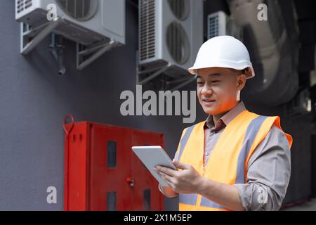 Smiling Asian man works in construction and manufacturing. Standing outside in a protective helmet and vest, holding a tablet, checking data, planning. Stock Photo