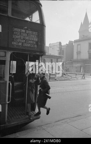 A DAY IN THE LIFE OF A WARTIME HOUSEWIFE: EVERYDAY LIFE IN LONDON, ENGLAND, 1941 - Mrs Day, helped by the female conductor, jumps on the bus that will take her to work. In the background, it is clear that quite a bit of air raid damage has been sustained. This photograph was probably taken on Fulham Road. The tower visible in the background is part of St Stephen's Hospital (now the Chelsea and Westminster Hospital), which was built in 1878 as the Fulham Workhouse and St George's Infirmary, Stock Photo