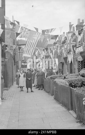 VE Day Scenes - A street scene. A group of people in the background of the image are looking towards the camera. Bunting and flags of the Allied nations are strung between the buildings. From up a tree to behind the camera.The image shows a dense crowd of people, gathered outside the gates of Buckingham Palace, on the 8th May, 1945 - VE-Day. The photograph was taken by Walter Lassally, a young man who had arrived in Britain, as a refugee six years earlier. Years later, Walter explained that as a child, growing up Berlin, he was told of a parade that a vast crowd had assembled to watch. Walter Stock Photo