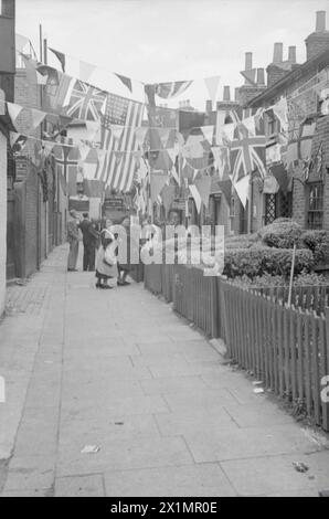 VE Day Scenes - A street scene. Three women, in the background of the image are looking towards the camera - two of the women are leaning on the garden fence, at the front of a house. Bunting and flags of the Allied nations are strung between the buildings. From up a tree to behind the camera.The image shows a dense crowd of people, gathered outside the gates of Buckingham Palace, on the 8th May, 1945 - VE-Day. The photograph was taken by Walter Lassally, a young man who had arrived in Britain, as a refugee six years earlier. Years later, Walter explained that as a child, growing up Berlin, he Stock Photo