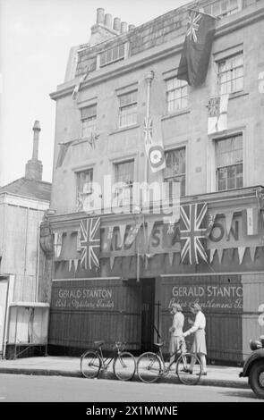 VE Day Scenes - A retail premises in Richmond, Surrey. The business bears the name Gerald Stanton, which has been largely obscured by bunting and flags - Union flags predominating. The shop windows are covered in protective hoarding, made from wooden planks. The name GERALD STANTON has been painted onto the hoardings. Two young women are outside the shop. Two women's bicycles are resting against the kerb. From up a tree to behind the camera.The image shows a dense crowd of people, gathered outside the gates of Buckingham Palace, on the 8th May, 1945 - VE-Day. The photograph was taken by Walter Stock Photo