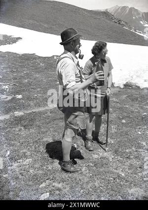 1950s, historical, standing on a high mountaint top, an swiss male alpine guide standing with a lady walker. The guide, wearing the traditional cothing of the era, leather breeches, hat and leather boots is smoking an alpine pipe and holding in his hand a bottle of wine. Stock Photo