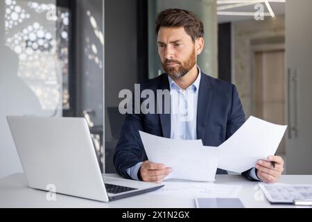 A serious young man businessman works in the office at a desk, looks at the screen of a laptop, holds documents in his hands, compares data, makes statistics and analysis. Stock Photo
