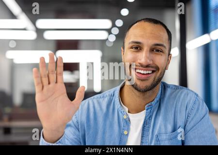 Close-up portrait of a young hispanic male office worker and freelancer sitting in front of the camera, greeting and waving at the camera. Stock Photo