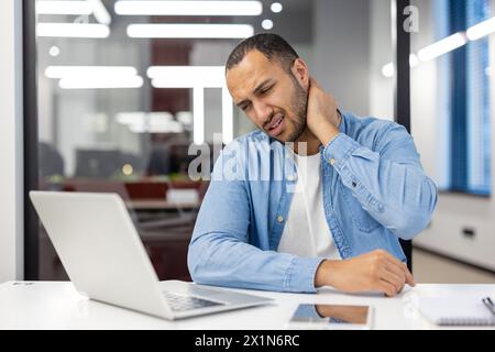 Tired hispanic man in shirt sitting in office at desk with laptop and holding hand on neck, doing massage, feeling pressure and pain. Stock Photo