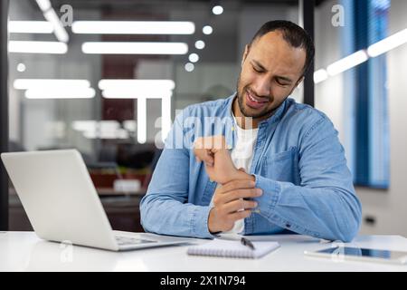 Muslim young man sitting at the office desk and holding his wrist with his hand, feeling pain and discomfort, having a massage and resting. Stock Photo