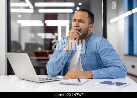 A tired young Muslim man in a blue shirt sits at a desk in a modern office and yawns while covering his mouth with his hand. Stock Photo