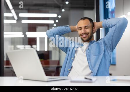 Smiling and relaxed young hispanic man sitting in the office at the desk, resting his hands behind his head and closing his eyes. Stock Photo