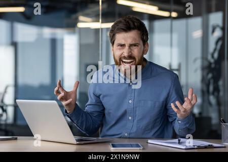 Portrait of an angry and irritated young businessman sitting at a desk in the office and shouting at the camera. Stock Photo