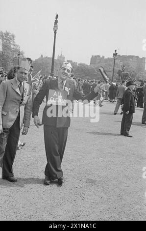 VE Day Scenes - A street scene in the area near The Tower of London. In the centre foreground a man wearing a suit and a party hat, is smiling directly into the camera. The man is waving a rattle in his left hand. there is a rosette on his left lapel. A sign hung from a loop of string around his neck reads 'ALL CLEAR'. There is a man adjacent to him, who is also wearing a fez party hat, with a checked jacket and chalk stripe trousers. This man has a more serious facial expression. From up a tree to behind the camera.The image shows people, gathered in the vicinity of The Tower of London, on th Stock Photo