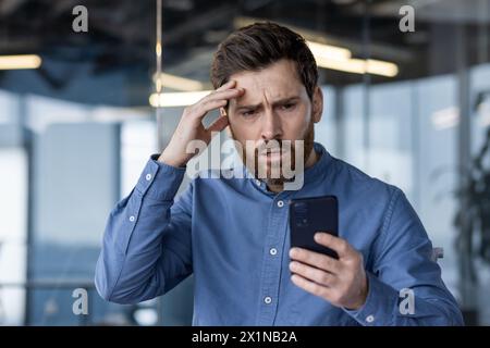Upset and serious young male businessman standing in office and looking shocked at phone screen, holding hand to head. Stock Photo