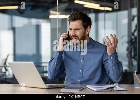 Angry and irritated young businessman sitting in the office at the desk and talking on the phone while waving his hands. Stock Photo