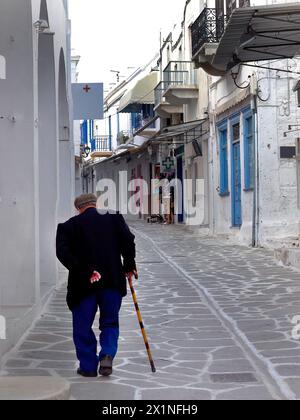 Backside of unrecognizable elderly senoir man wearing beret hat holding a cane walking in the small mediterranean alley of Paros island, local life Stock Photo