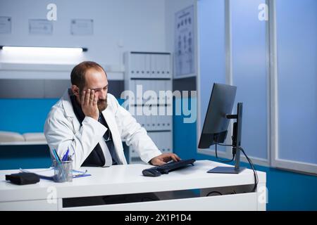 Image showcasing a tired caucasian doctor seated at a desk with a desktop computer after a long day in hospital. Exhausted male physician touching his face in a clinic office room. Stock Photo