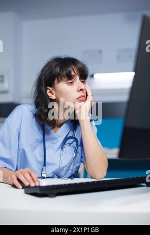 Caucasian female practitioner at a modern clinic desk, tired but focused. Before an appointment, pharmacist trained nurse analyzes a medical checkup using computer in the hospital office. Stock Photo