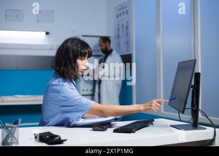 Image shows a nurse practitioner at the clinic desk reviewing and analyzing her notes. A Caucasian woman in blue scrubs prepares for patient medical consultations by using a desktop pc and clipboard. Stock Photo