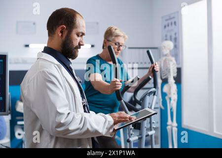 Portrait of doctor holding tablet while elderly patient uses electronic bicycle for physical rehabilitation. Male medical physician in lab coat using digital device for medical physiotherapy research. Stock Photo