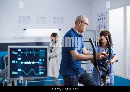 Nurse practitioner giving assistance to old man using stationary bike for physical activity and recovery. Medical assistant helping male patient with exercise bicycle for physiotherapy. Stock Photo