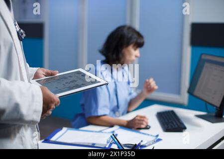 Close-up shot of medical professionals utilizing modern technology for discussing healthcare in a clinic. Selective focus shows a person holding a tablet and a female nurse using a computer. Stock Photo