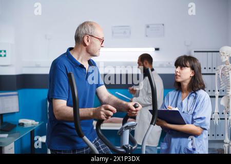 Elderly patient is seen receiving physiotherapy on stationary bike, assisted by female nurse dressed in blue scrubs. Senior man is getting rehabilitative care with help from health professional. Stock Photo