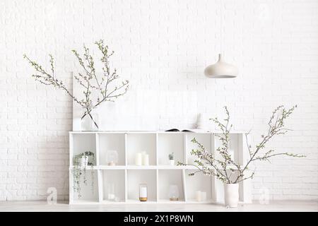 Vases with blooming branches on shelving unit near white brick wall Stock Photo