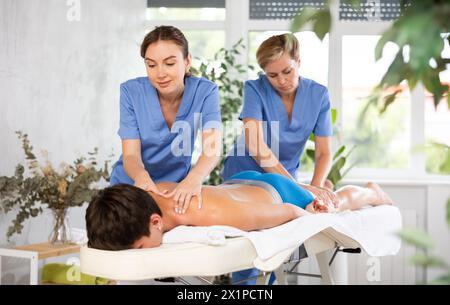 Two female masseuses perform stroking and rubbing of muscles of back and shoulders to guy client Stock Photo