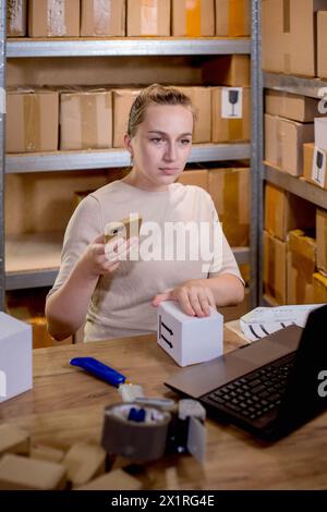 Female small business owner using mobile app on smartphone checking parcel box. Warehouse worker, seller holding phone scanning retail dropshipping pa Stock Photo