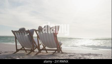 Senior biracial couple relaxing on beach chairs by sea Stock Photo