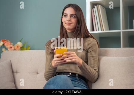 Smiling young woman sitting on a sofa looking at social media with a smartphone Stock Photo