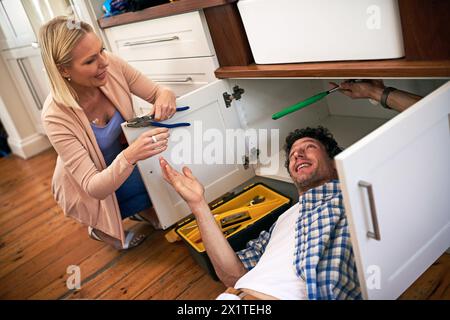 Lady, help and handyman with tools for fixing home basin, drain or broken interior sewer. Plumber, working and woman watching male person repair for Stock Photo