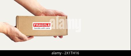 One rectangular cardboard box in hands for fragile items packing on a plain light background. Red warning label, sticker for packaging 'Fragile' Stock Photo