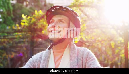 Cycling, face and helmet with senior woman outdoor in garden for retirement recreation or wellness. Exercise, smile and thinking with happy cyclist at Stock Photo