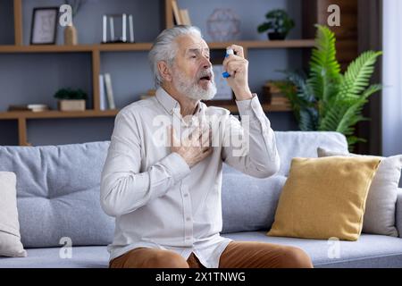 Elderly Caucasian man experiencing an asthma attack, using a blue inhaler while sitting on a couch in a modern living room. Stock Photo