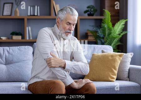 An elderly man experiencing discomfort holds his shoulder while seated on a couch, illustrating health issues in senior living. Stock Photo