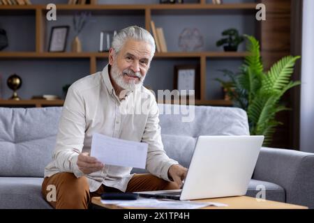 A cheerful elderly man works from his cozy living room, using a laptop and holding documents. He smiles warmly, expressing contentment and productivity while working remotely. Stock Photo