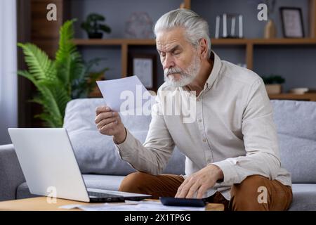 Focused senior man with gray beard sitting on a sofa, examining papers while working on a laptop in a modern home office setting. Stock Photo