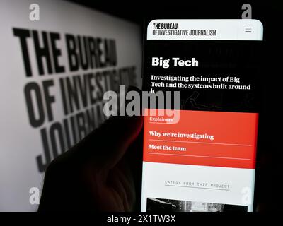 Person holding cellphone with webpage of organisation The Bureau of Investigative Journalism (TBIJ) with logo. Focus on center of phone display. Stock Photo