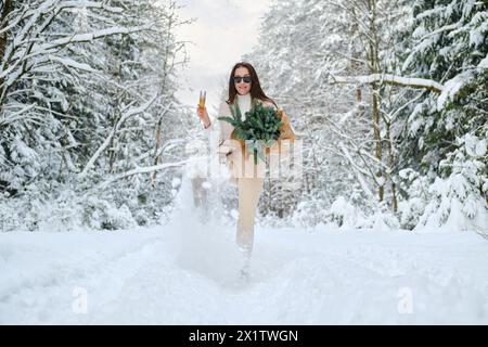 Playful woman kicking snow in winter forest holding glass of champagne and fir branches in hand Stock Photo