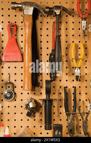 Assorted hand tools that include a small and medium hammer, red needle nose pliers, yellow wire cutter, standard black pliers on pegboard wall in Stock Photo
